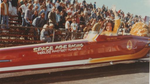 Smiling woman sits in a racecar and waves to the camera. In the bleachers behind her, spectators watch.