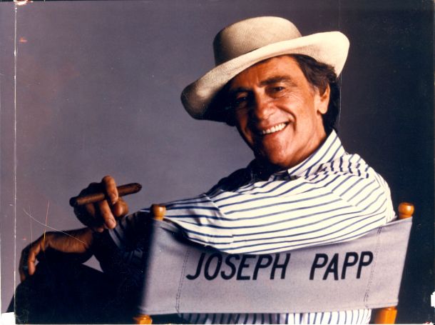 Theater producer Joe Papp sitting in director's chair with his name on it and holding a cigar.